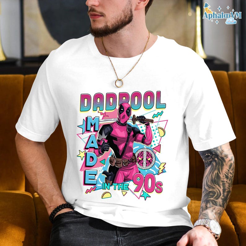 A white t-shirt with a graphic design featuring a character from the Marvel Cinematic Universe, specifically the character Deadpool. The design includes the text "DAD POOL MADE IN THE 90'S" and includes various colorful elements and symbols associated with the character.