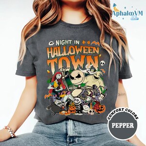 May include: A charcoal gray short-sleeve t-shirt featuring a graphic print with the text NIGHT IN HALLOWEEN TOWN. The design includes various animated characters, pumpkins, and bats in orange, white, and green colors. The shirt has a crew neckline.
