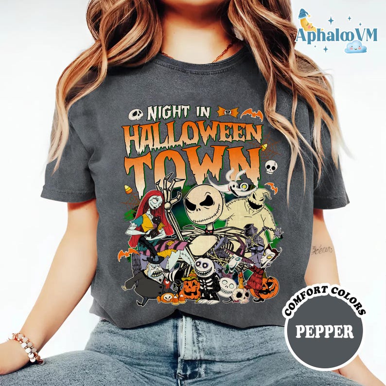 May include: A charcoal gray short-sleeve t-shirt featuring a graphic print with the text NIGHT IN HALLOWEEN TOWN. The design includes various animated characters, pumpkins, and bats in orange, white, and green colors. The shirt has a crew neckline.