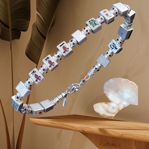 May include: Silver bracelet featuring square charms with Chinese characters and red and green accents. The bracelet is connected with a silver chain and clasp. The text "Now in Silver!" is at the bottom of the image.