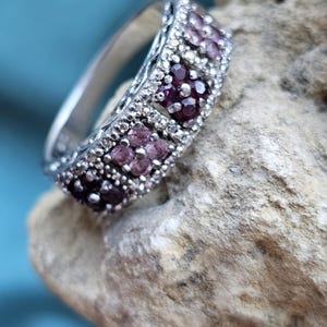 May include: A silver-toned ring featuring a band with a textured, granulated surface. The ring is set with multiple small, round, deep red and light pink gemstones arranged in floral-like clusters along the top of the band.