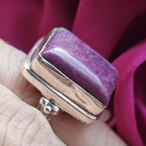 May include: A silver ring featuring a large, rectangular, polished purple gemstone set in a smooth metal bezel. The stone has a deep magenta hue with subtle natural inclusions and a glossy surface. The band is made of polished silver metal.