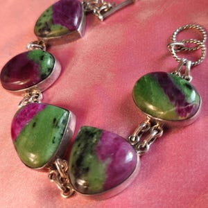 May include: A silver bracelet with oval gemstones in shades of green and deep red. The stones are set in silver and linked together. The bracelet is secured with a toggle clasp.