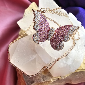 May include: A rose gold necklace featuring a butterfly pendant encrusted with small pink and clear cubic zirconia stones. The butterfly wings display a gradient pattern of pink stones in the center and clear stones along the outer edges.