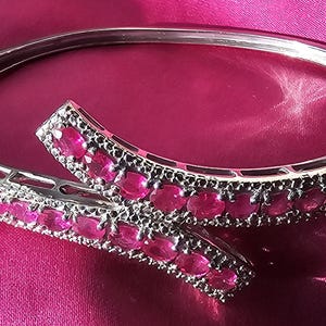 Puede incluir: Un brazalete rígido articulado de tono plateado con un diseño de bypass y dos extremos superpuestos. El brazalete presenta una hilera continua de piedras ovaladas de color rosa vibrante en la parte superior, con pequeñas piedras claras en los bordes.