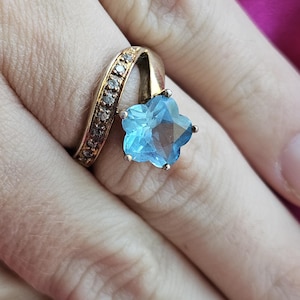May include: A gold-toned ring featuring a prominent star-shaped light blue gemstone as the centerpiece. The band is curved and embellished with a row of small, clear, sparkling stones along the side, creating a distinct and elegant appearance.