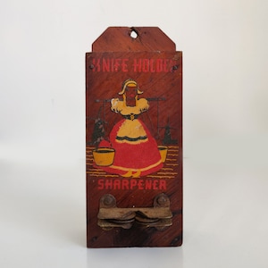Op de afbeelding: Vintage houten messenhouder en slijper met een geschilderd beeld van een vrouw in een rode en gele jurk. De tekst "KNIFE HOLDER SHARPENER" is op de voorkant gedrukt.