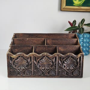 May include: A dark brown wooden desk organizer with multiple compartments. The front features carved floral designs. The organizer has a distressed finish, giving it a vintage appearance. It is designed to hold letters, documents, or office supplies.