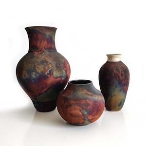 Puede incluir: Tres jarrones de cerámica con acabado mate que presentan un patrón arremolinado y moteado de rojo intenso, naranja quemado, marrón oscuro y toques de azul verdoso metálico. Los recipientes varían en tamaño y forma, con uno alto, uno mediano y un cuenco.