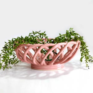 May include: A light pink ceramic bowl featuring a woven, lattice-style design with a circular base. The bowl is filled with a trailing green plant that has small, round leaves cascading over the sides of the open, interconnected ceramic structure.