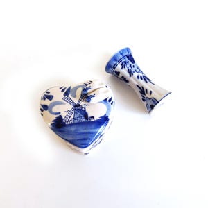 May include: Two ceramic pieces with a blue and white windmill design. One is heart-shaped with a lid, the other is a small vase. The pieces have a traditional Delftware style, with a blue and white color scheme.