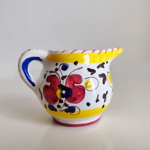 May include: A small, white ceramic creamer with a handle and spout. The creamer is decorated with red floral designs, blue accents, and yellow trim. The handle is blue, and the rim is yellow with red stripes.