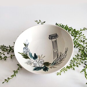 May include: A white ceramic bowl featuring a black line drawing of a classical column on the interior. The bowl is decorated with hand-painted floral motifs in shades of green and blue, arranged in a curved pattern around the base of the column.