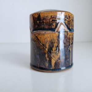 May include: A cylindrical ceramic container with a lid. The container is glazed in shades of brown, tan, and dark blue, with a textured, crackled appearance. The lid fits snugly on top. The handmade pottery piece is likely for storage.