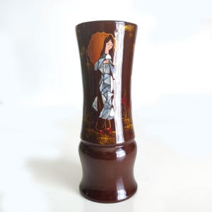 May include: A dark brown vase with a decorative design. The vase features a woman with an umbrella, depicted in a mosaic style. The woman is wearing a blue dress and red shoes. Gold accents and floral details are also present.