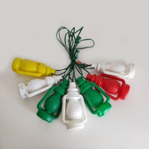Puede incluir: Una cadena de luces navideñas con forma de linternas de colores. Las linternas son de varios colores, incluyendo amarillo, blanco, verde y rojo. Las luces están conectadas por un cable verde y dispuestas en forma circular.