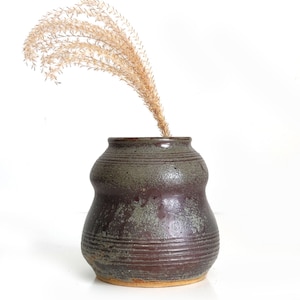 May include: A dark brown ceramic vase with a textured, mottled glaze and a rounded, bulbous shape. The vase has a wide opening and is decorated with horizontal lines. A dried grass stalk is in the vase.