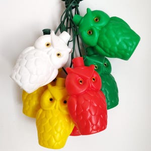 May include: A string of owl-shaped Christmas lights in white, yellow, red, and green. Each owl has a textured surface and golden-yellow eyes. The lights are strung on a green wire, creating a festive and whimsical decoration.