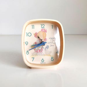 May include: A vintage, cream-colored alarm clock with a square face. The clock features a cartoon illustration of a girl and the text "Waddle I Do Without You." The clock hands are blue, and the numbers are in a teal color. The clock is marked "MADE IN USA."