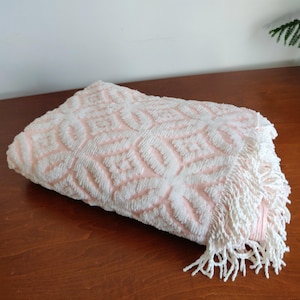 May include: A folded pink and white patterned towel with a fringe. The towel features a geometric design in white against a pink backdrop. The fringe is white and hangs from one edge of the towel.