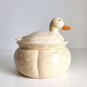 May include: A ceramic lidded jar featuring a sculpted duck sitting atop a base textured to resemble a woven basket. The duck is off-white with an orange beak, while the round base has a light tan finish with a detailed basketweave pattern.