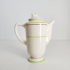 May include: Cream-colored ceramic coffee pot with a lid and handle. The pot is adorned with horizontal green and brown stripes. The base and lid are also accented with green trim. The spout is on the right side.