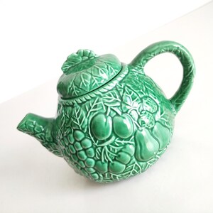 Puede incluir: Una tetera de cerámica de color verde brillante con un patrón en relieve detallado de varias frutas y hojas que cubre toda la superficie exterior, incluyendo una tapa a juego con un asa en forma de hoja y un pico curvo.