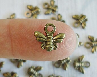 May include: Bronze-colored bee charms with a loop for attaching to jewelry or crafts. The bee charms are small, detailed, and have a vintage aesthetic. Several charms are scattered around the main charm, which is held by a finger.