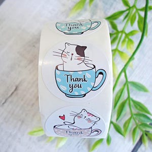 May include: A roll of white, round stickers featuring a cartoon cat in a teacup. The cat is white with a brown patch. The teacup is light blue with the words "Thank you" on it. A green plant is in the background.