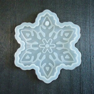 May include: A translucent, flexible silicone mold shaped like a six-pointed snowflake. The mold features a raised, intricate snowflake pattern with a small circular indentation at the top point, designed for creating hanging ornaments or decorations.