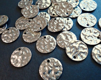 May include: A pile of silver-toned metal round discs with a hammered texture and two holes. The discs are scattered on a dark blue surface.