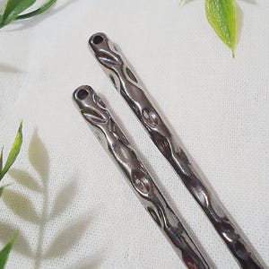 Pu&ograve; includere: Due forcine per capelli argentate con una texture martellata. Le forcine hanno una parte superiore arrotondata con un piccolo foro e un design ondulato e strutturato lungo la lunghezza. Lo sfondo &egrave; bianco con foglie verdi.