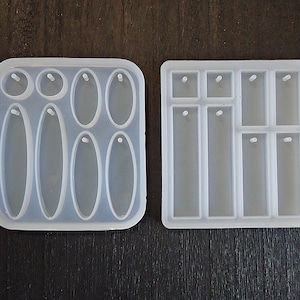 May include: Two clear silicone molds for crafting. One mold features various oval and circular shapes with holes for jewelry making. The other mold has rectangular shapes, also with holes, for creating pendants or charms. Both molds are on a dark surface.