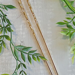 Pu&ograve; includere: Due forcine per capelli color oro con un design strutturato e accenti rossi sulla parte superiore. Le forcine sono posizionate in diagonale su una superficie di legno chiaro, con viti verdi sullo sfondo.