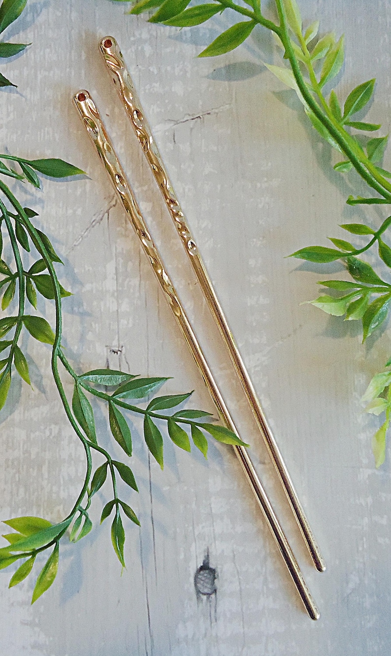 Pu&ograve; includere: Due forcine per capelli color oro con un design strutturato e accenti rossi sulla parte superiore. Le forcine sono posizionate in diagonale su una superficie di legno chiaro, con viti verdi sullo sfondo.