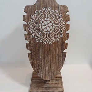 May include: A wooden jewelry display stand with a distressed finish. The stand features a decorative white mandala design and multiple slots for necklaces. The base is square and the overall height is approximately 12 inches.