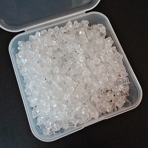 May include: A clear, plastic container filled with numerous clear earring backs. The earring backs are small, with a variety of shapes, and appear to be made of a soft, flexible material. The container is open, revealing the contents.