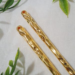 Pu&ograve; includere: Due forcine per capelli color oro con un design ornato. Ogni forcina ha una piccola gemma rotonda di colore arancione sulla parte superiore. Le forcine sono appoggiate su una superficie bianca e strutturata, con foglie verdi sullo sfondo.