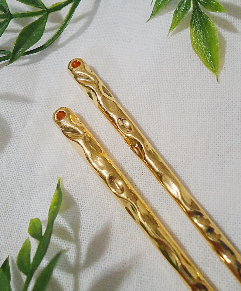 Pu&ograve; includere: Due forcine per capelli color oro con un design ornato. Ogni forcina ha una piccola gemma rotonda di colore arancione sulla parte superiore. Le forcine sono appoggiate su una superficie bianca e strutturata, con foglie verdi sullo sfondo.