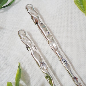 Pu&ograve; includere: Due forcine per capelli color argento con un design martellato e strutturato. Le forcine hanno un aspetto attorcigliato e sono poste su uno sfondo bianco con accenti di fogliame verde, creando un'estetica naturale.