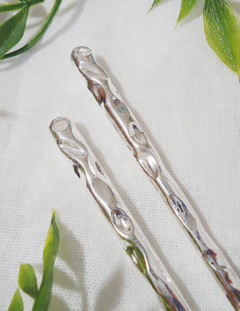 Pu&ograve; includere: Due forcine per capelli color argento con un design martellato e strutturato. Le forcine hanno un aspetto attorcigliato e sono poste su uno sfondo bianco con accenti di fogliame verde, creando un'estetica naturale.