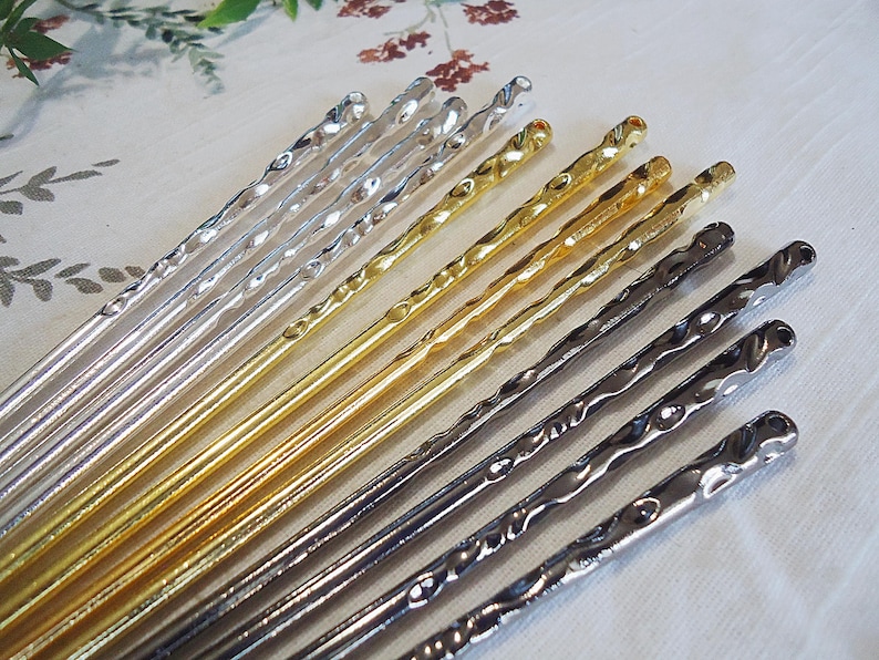 Pu&ograve; includere: Un assortimento di bastoncini decorativi per capelli in argento, oro e grigio scuro. Ogni bastoncino presenta un design a spirale lungo la sua lunghezza e una parte superiore arrotondata. I bastoncini sono disposti su una superficie bianca, offrendo una variet&agrave; di tonalit&agrave; metalliche.