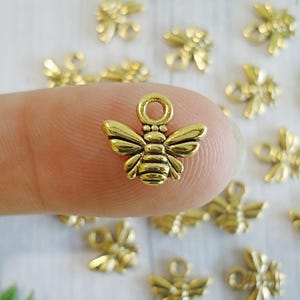May include: Close-up of several small, gold-colored bee charms. The bee charms have detailed wings, bodies, and a loop at the top for attaching to jewelry or other crafts. The background is a light color.