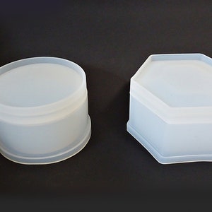 May include: Two white plastic containers, one round and one hexagonal. Both containers have lids.
