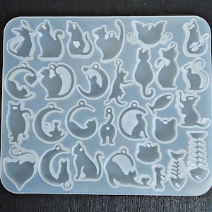 May include: A clear silicone mold filled with various cat-themed shapes. The mold includes cat silhouettes, fish, moons, and bone shapes. The shapes are designed for crafting and creating resin or other decorative items. The mold is rectangular with rounded corners.