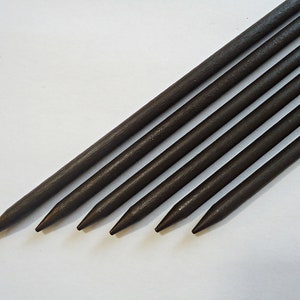 May include: A collection of dark brown wooden skewers, each with a sharpened point. The skewers are arranged diagonally across a white surface, showcasing their uniform shape and color. The image highlights the natural wood grain.