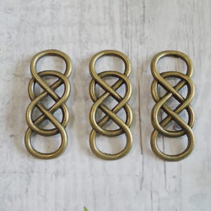 May include: Three bronze-colored infinity knot charms. Each charm features a figure-eight design with interlocking loops. The charms are made of metal and have a slightly aged appearance, suggesting a vintage or antique style. They are suitable for jewelry making or craft projects.