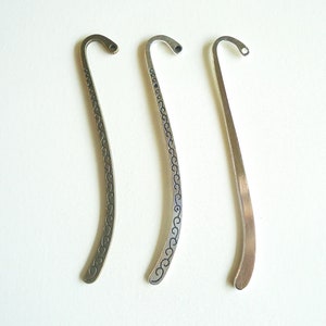 May include: Three silver-toned metal bookmarks with curved tops and decorative designs. The bookmarks are arranged on a white surface. The two left bookmarks have etched patterns, while the right one is plain.