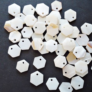 May include: A collection of white, iridescent, hexagonal shell charms with a hole in the center. The charms are scattered on a black surface.