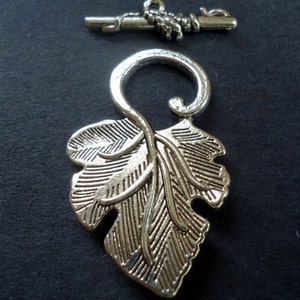 May include: Silver tone leaf-shaped toggle clasp with a decorative swirl design. The clasp is attached to a silver tone bar with a decorative knot.